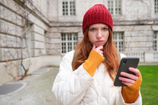 Portrait d'une jeune femme rousse au chapeau rouge tient un smartphone et a l'air pensif en fronçant les sourcils avec perpl