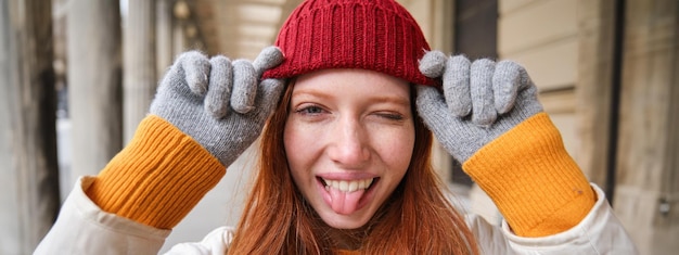 Photo gratuite portrait d'une jeune femme rousse en bonnet et gants tricotés sourit et regarde de côté se promène dans la ville