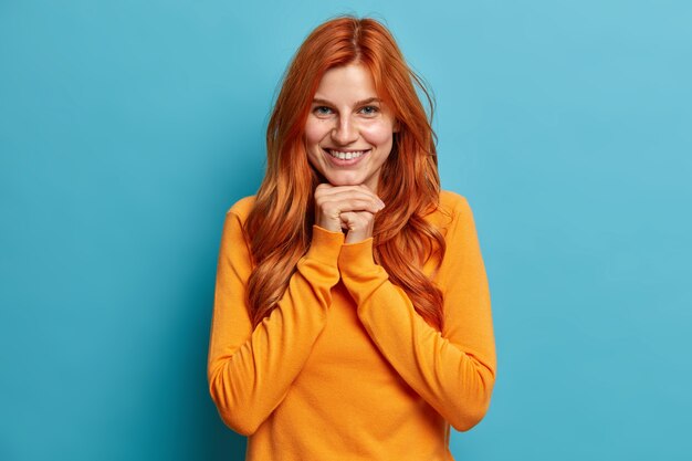 Portrait de jeune femme rousse à la recherche agréable sourit joyeusement et garde les mains sous le menton regarde directement avec une expression satisfaite vêtue de vêtements décontractés.