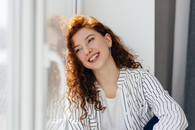 Portrait de jeune femme rousse souriante assise sur le rebord de la fenêtre