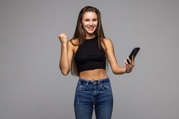 Portrait d'une jeune femme satisfaite tenant un téléphone portable et célébrant isolé sur fond gris