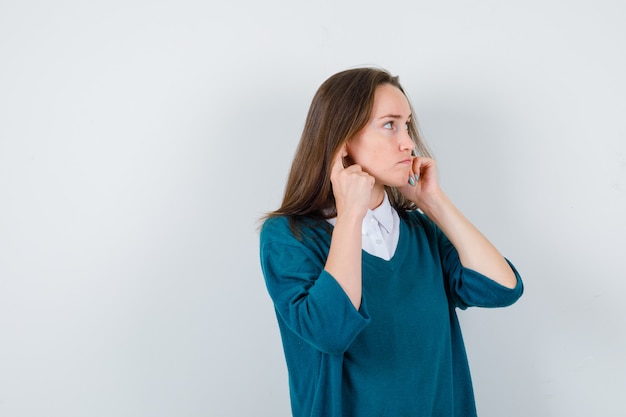 Photo gratuite portrait de jeune femme se branchant les oreilles avec les doigts, regardant de côté en chemise blanche, pull et regardant la vue de face focalisée