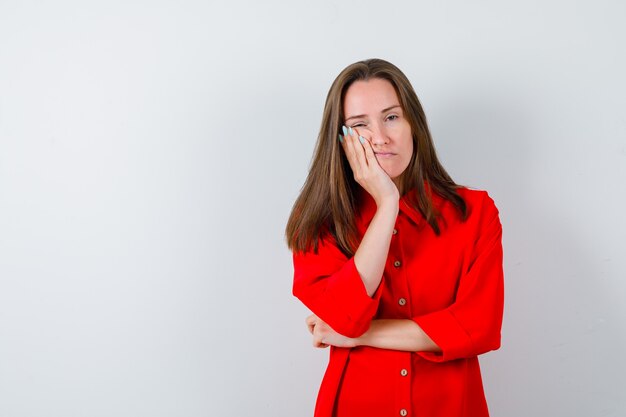 Portrait de jeune femme se penchant la joue sur la paume en chemisier rouge et à la vue de face ennuyée