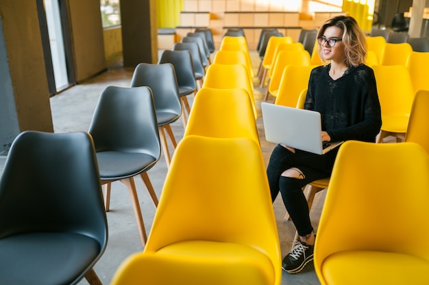 Portrait de jeune femme séduisante assise dans une salle de conférence travaillant sur un ordinateur portable portant des lunettes, l'apprentissage des élèves en classe avec de nombreuses chaises jaunes