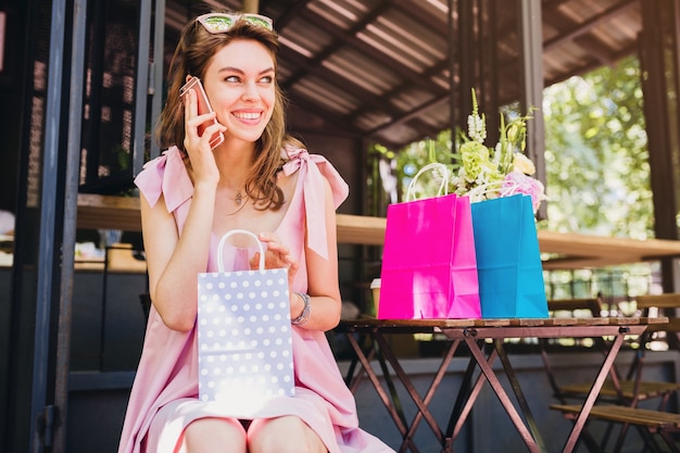 Portrait de jeune femme séduisante heureuse souriante assise dans un café, parler au téléphone avec des sacs à provisions, tenue de mode d'été, style hipster, robe en coton rose, vêtements à la mode