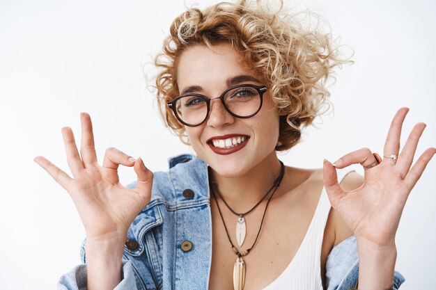 Portrait d'une jeune femme séduisante, joyeuse et charismatique, optimiste et charismatique, aux cheveux blonds et aux lunettes, souriant joyeusement montrant un geste correct recommandant une excellente application, donnant son approbation