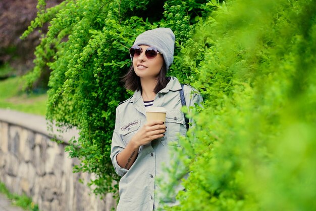 Portrait de jeune femme séduisante à lunettes de soleil tient une tasse de café en papier dans un parc d'été verdoyant.