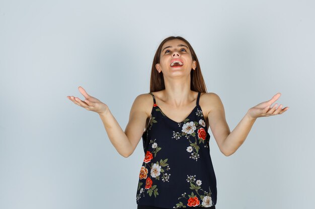 Portrait d'une jeune femme séduisante soulevant des paumes écartées tout en levant les yeux en blouse et en regardant la vue de face reconnaissante