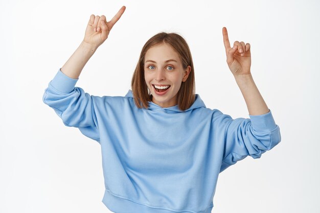 Portrait d'une jeune femme séduisante souriante, pointant du doigt le logo, montrant une bannière de réduction de vente, une publicité vers le haut, debout dans un sweat à capuche bleu, recommande de cliquer sur le lien, fond blanc.