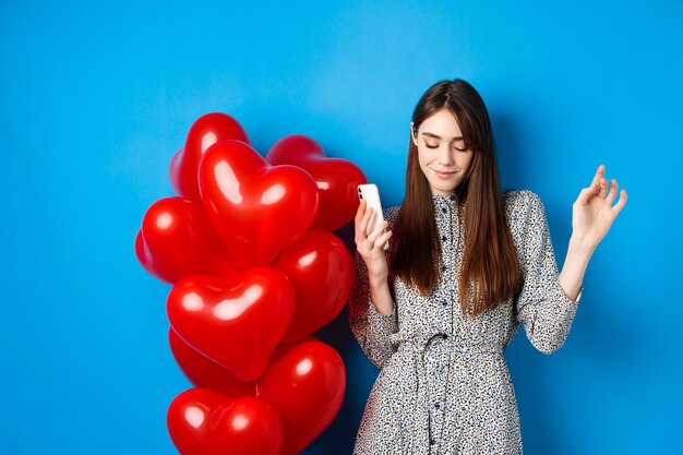 Portrait d'une jeune femme séduisante tenant un smartphone et dansant près de ballons rouges de la Saint-Valentin, debout sur fond bleu