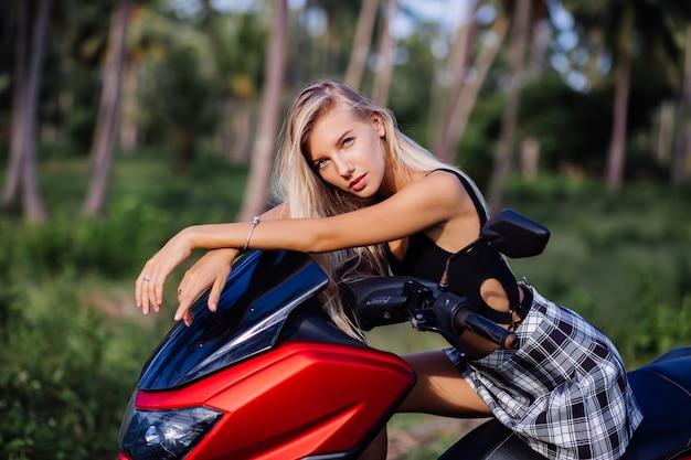 Portrait de jeune femme slim fit caucasien européen en maillot de bain haut noir et jupe écolière à carreaux noir et blanc sur le champ de la jungle tropicale avec moto, moto. Lunettes de soleil et casquette noire