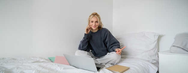 Portrait d'une jeune femme souriante étudiant dans son lit travaillant à domicile dans la chambre à coucher assise avec un ordinateur portable