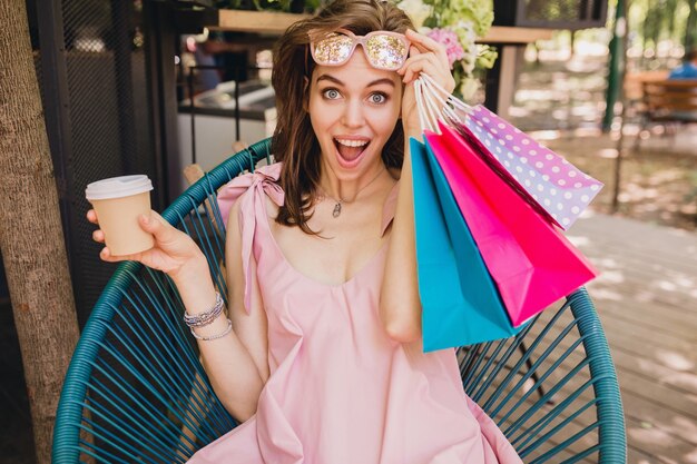 Portrait d'une jeune femme souriante et heureuse avec une expression de visage excitée assise dans un café avec des sacs à provisions en train de boire du café