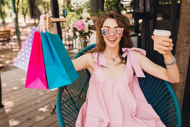 Portrait de jeune femme souriante heureuse jolie avec l'expression du visage excité assis dans un café avec des sacs à provisions boire du café, tenue de mode d'été, robe en coton rose, vêtements à la mode