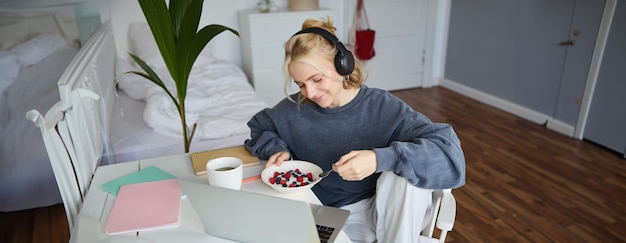 Photo gratuite portrait d'une jeune femme souriante regardant une émission de télévision avec des écouteurs, prenant son petit déjeuner et regardant