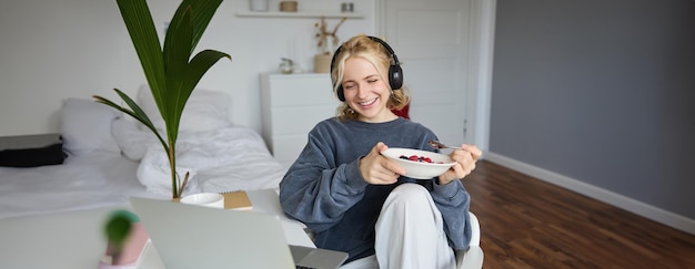 Portrait d'une jeune femme souriante regardant une émission de télévision avec des écouteurs, prenant son petit déjeuner et regardant