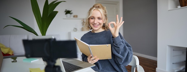 Photo gratuite portrait d'une jeune femme souriante tenant un bloc-notes montrant un signe ok ok en regardant un appareil photo numérique