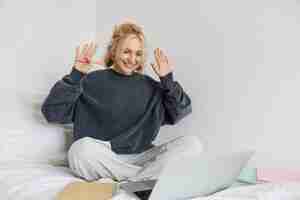 Photo gratuite portrait d'une jeune femme souriante travaillant depuis son lit assise dans la chambre à coucher avec un ordinateur portable et des documents