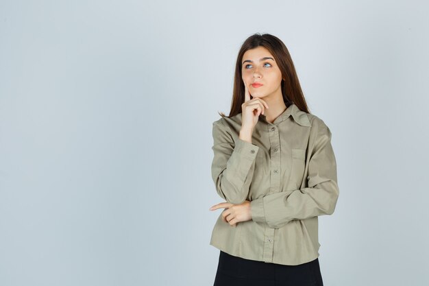 Portrait de jeune femme soutenant le menton à portée de main, tenant le doigt sur la joue en chemise, jupe et regardant pensive vue de face