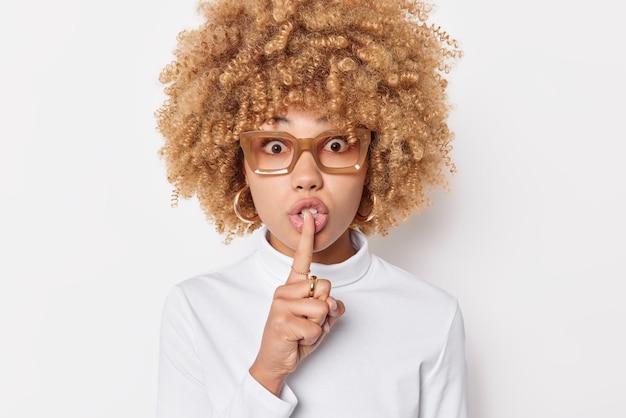 Portrait d'une jeune femme surprise aux cheveux bouclés fait un geste de silence garde l'index sur les lèvres exige un silence complet porte des lunettes et un pull décontracté isolé sur fond blanc.