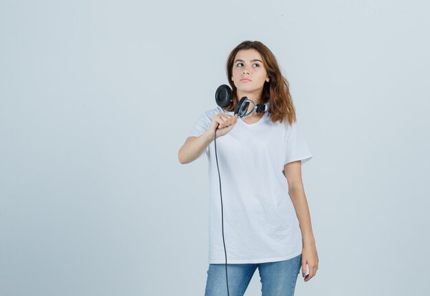 Portrait de jeune femme tenant un casque tout en pensant en t-shirt blanc, jeans et à la charmante vue de face