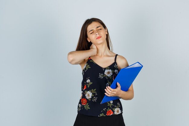 Portrait de jeune femme tenant un dossier, ayant des douleurs au cou en chemisier, jupe et à la vue de face fatiguée