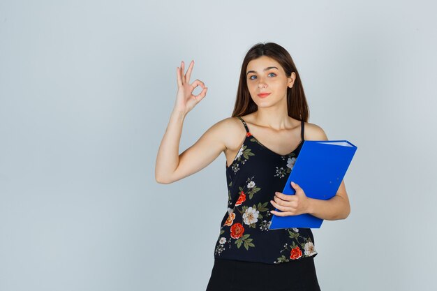 Portrait de jeune femme tenant un dossier tout en montrant un geste correct en chemisier, jupe et regardant la vue de face confiante