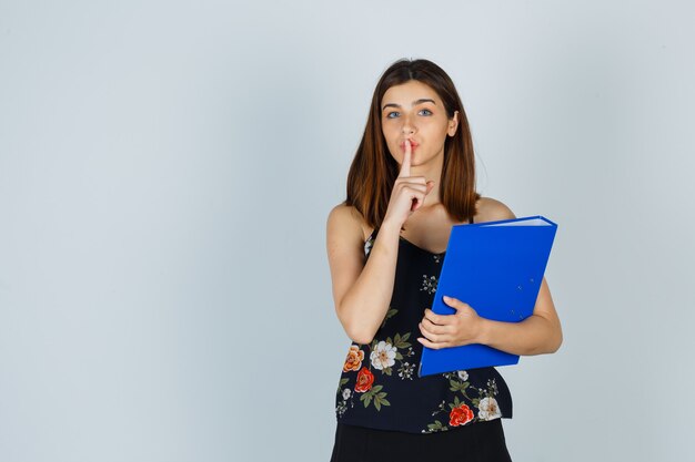 Portrait de jeune femme tenant un dossier tout en montrant un geste de silence en blouse, jupe et en regardant attentivement la vue de face