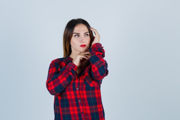 Photo gratuite portrait de jeune femme touchant la tête, soutenant le menton sur la main, regardant loin en chemise à carreaux et regardant pensive vue de face