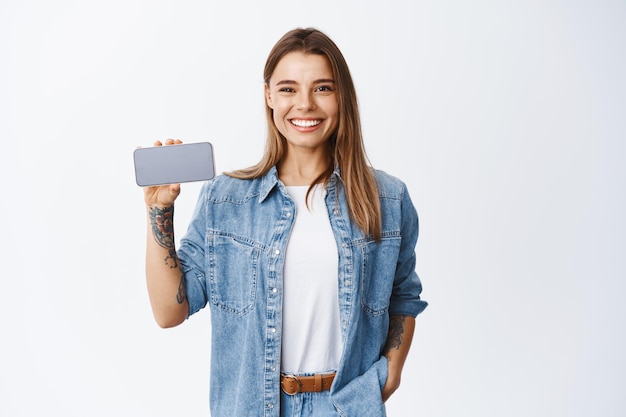 Portrait de jeune femme en vêtements décontractés tenant un smartphone à l'horizontale, montrant un écran de téléphone portable vide pour la publicité de l'application, blanc