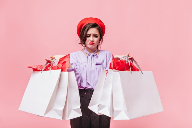 Portrait de jeune fille au béret rouge à la mécontentement des paquets avec des vêtements. Dame en chemisier lilas et pantalon noir posant sur fond rose.