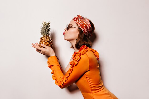 Portrait de jeune fille aux cheveux courts en bandeau élégant et robe orange embrassant l'ananas sur l'espace blanc.