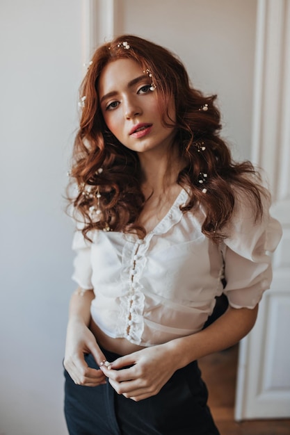 Portrait de jeune fille aux cheveux ondulés rouges vêtue d'un pantalon noir et d'un chemisier blanc Femme romantique avec des fleurs dans les cheveux posant dans la salle blanche