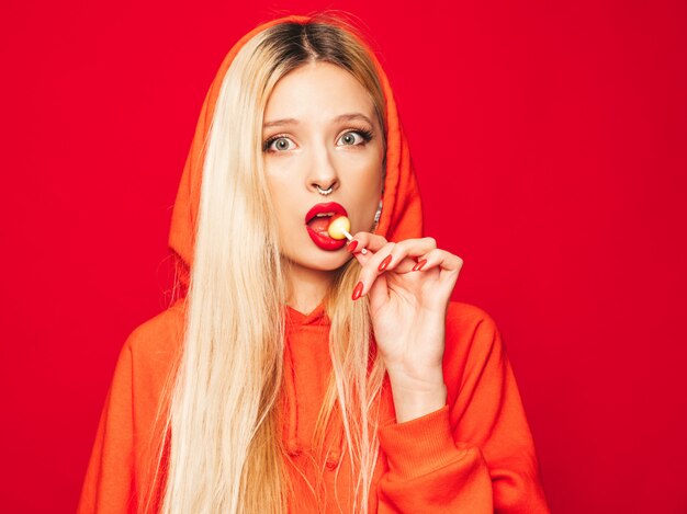 Portrait de jeune fille belle hipster belle mauvaise à capuche rouge à la mode et boucle d'oreille dans son nez. Modèle positif léchant des bonbons de sucre ronds