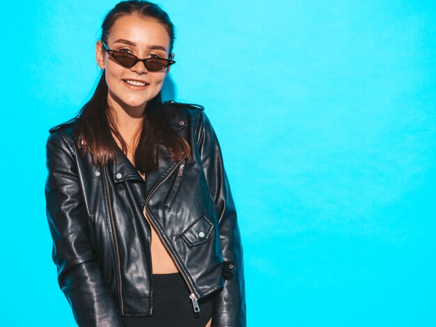 Portrait de jeune fille belle hipster belle mauvaise en veste de cuir noir tendance été et lunettes de soleil. Sexy femme insouciante isolée sur bleu. Brunette modèle posant