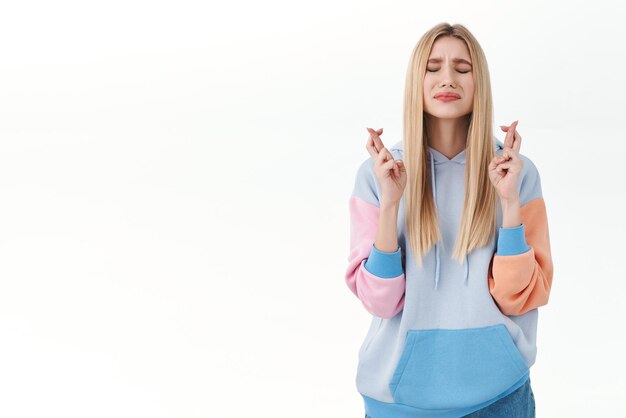 Portrait d'une jeune fille blonde pleine d'espoir aux cheveux longs, aux yeux fermés et aux doigts croisés boudeurs, bonne chance