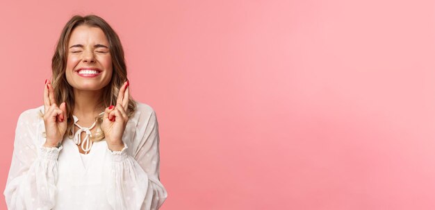 Portrait d'une jeune fille blonde pleine d'espoir excitée faisant un souhait croisé les doigts pour la bonne chance ferme les yeux et souriant en mettant tout en œuvre pour prier en implorant que le rêve devienne réalité en anticipant sur fond rose