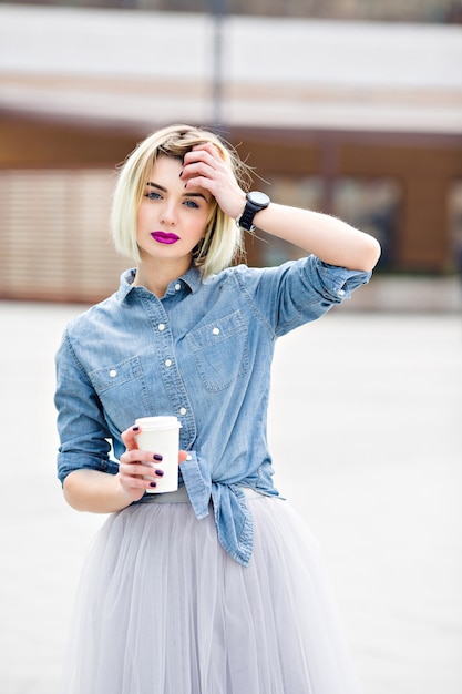 Un portrait d'une jeune fille blonde rêveuse debout avec des lèvres rose vif hoding une tasse de café et tenant une main près de sa tête