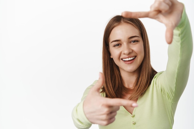 Portrait d'une jeune fille blonde souriante regarde à travers des cadres de doigts créatifs, riant, imaginant qch, faisant une prise de vue, debout heureux sur fond blanc