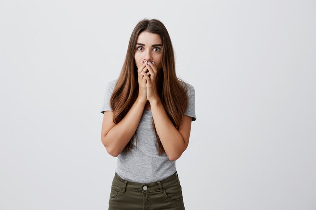 Portrait de jeune fille brune caucasienne magnifique aux cheveux longs dans des vêtements gris décontractés, fermant la bouche avec les mains avec une expression choquée et effrayée.