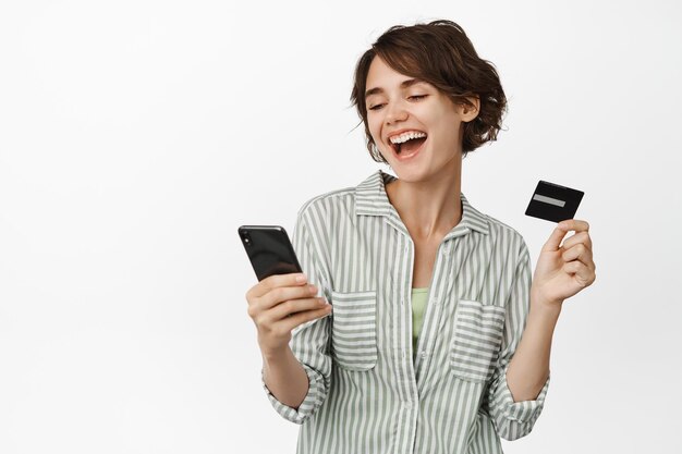 Portrait de jeune fille brune payant, shopping en ligne avec téléphone portable et carte de crédit, achat dans l'application, debout sur fond blanc. Copier l'espace