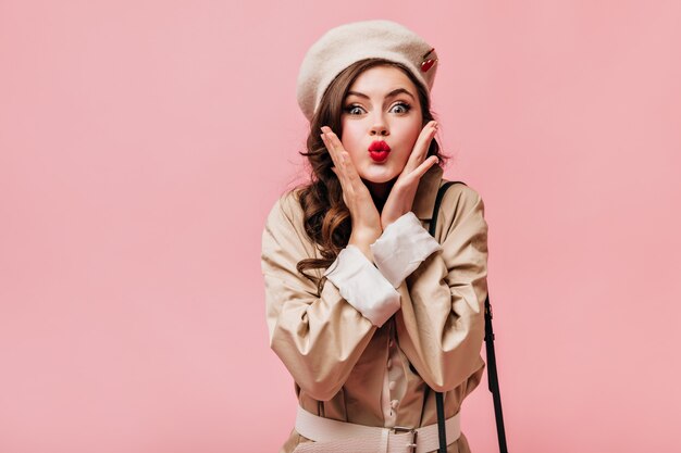 Portrait de jeune fille brune soufflant baiser. Dame aux yeux verts en béret et trench-coat posant sur fond rose.