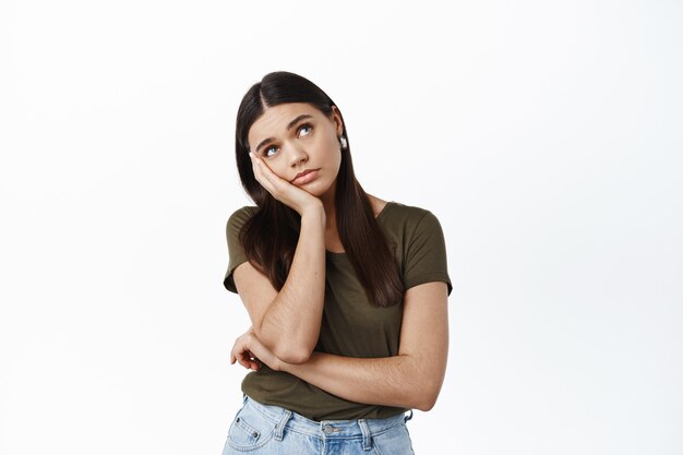 Portrait d'une jeune fille brune triste et ennuyée la tête penchée et regarde le coin supérieur droit pensif, n'a rien à faire, assiste à une réunion ennuyeuse, debout sur un mur blanc