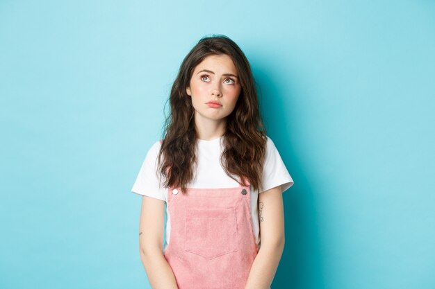 Portrait d'une jeune fille brune triste et sombre avec un maquillage glamour, regardant le coin supérieur gauche bouleversé et réfléchi, se sentant de mauvaise humeur, debout sur fond bleu.