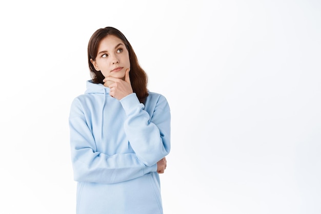 Portrait de jeune fille brune avec un visage pensif touchant le menton et regardant de côté le coin supérieur droit faisant des achats de choix et pensant debout sur fond blanc