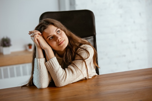 Photo gratuite portrait de jeune fille caucasienne dans des vêtements décontractés semble rêveur, mignon et heureux. levant les yeux et réfléchissant, assis à l'intérieur à la table en bois. concept d'avenir, cible, rêves, visualisation.