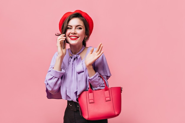 Portrait de jeune fille en chemisier lilas et béret rouge sur fond rose. Femme tenant un petit sac et parler au téléphone.