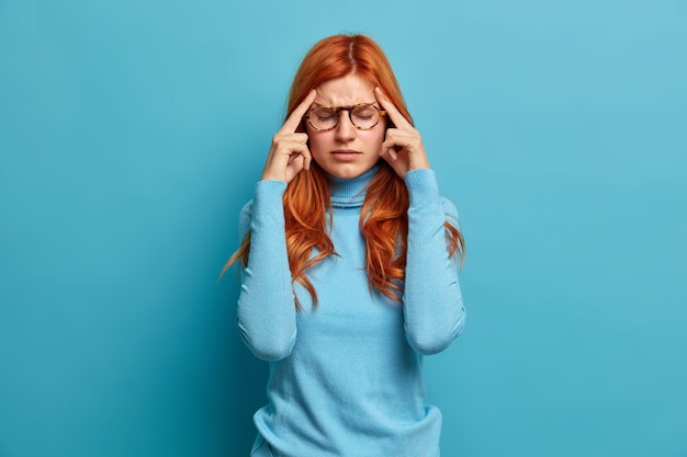 Portrait de jeune fille européenne au gingembre souffre de maux de tête sévères garde l'index sur les tempes tente de se concentrer et de continuer à travailler habillé de vêtements décontractés.