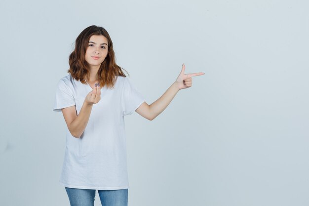 Portrait de jeune fille faisant un geste italien, pointant vers le côté droit en t-shirt blanc, jeans et à la vue de face confiante