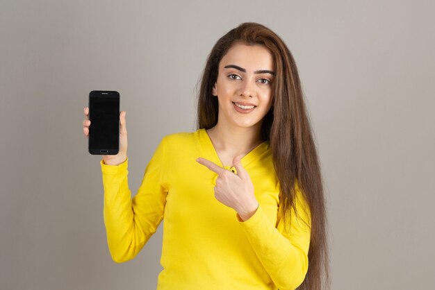 Portrait de jeune fille en haut jaune posant avec téléphone portable sur mur gris.